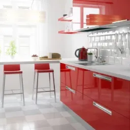 Jasna kuchnia z błyszczącymi, czerwonymi frontami szafek, białym blatem i białymi płytkami na ścianie, uzupełniona o srebrne akcenty i wysoki stół z czerwonymi stołkami.