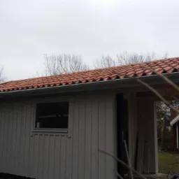 Mały, szary budynek gospodarczy z nową, czerwoną dachówką ceramiczną i szarymi rynnami, widok z zewnątrz, w tle drzewa bez liści i fragment innego budynku.