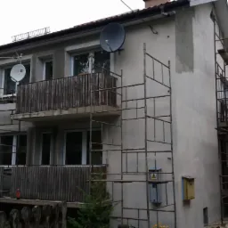 Dwupiętrowy dom w trakcie prac elewacyjnych, otoczony metalowymi rusztowaniami, z widocznymi satelitami na balkonach i świeżo otynkowaną fasadą.