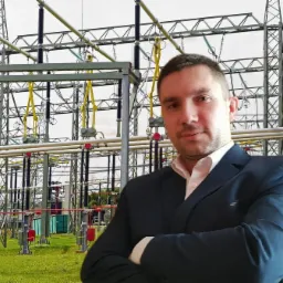 Portret mężczyzny w garniturze na tle stacji transformatorowej z widocznymi przewodami i konstrukcjami wsporczymi.