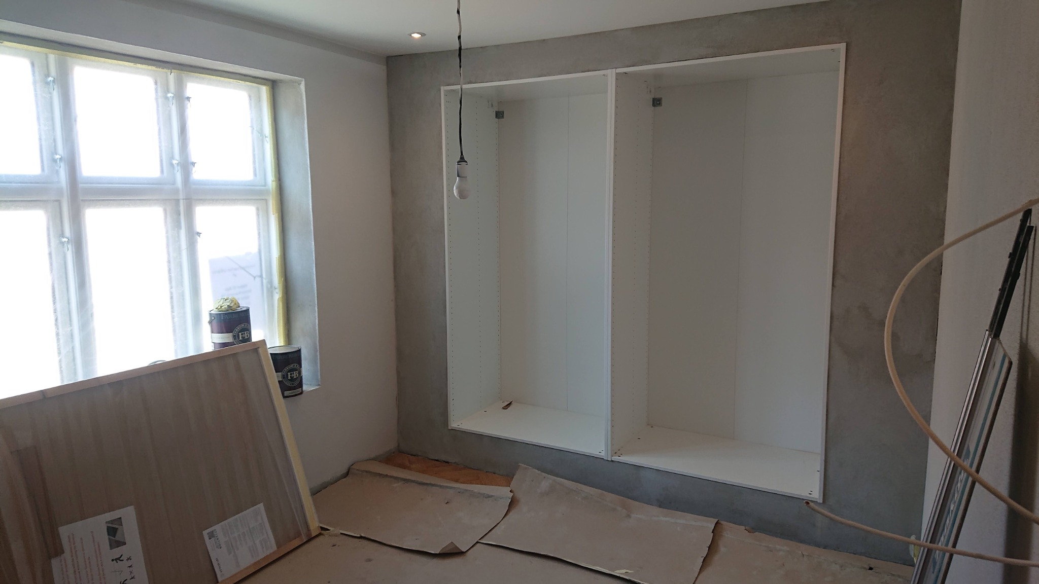 Wnętrze pokoju w trakcie prac wykończeniowych: surowe ściany, wnęka z białymi półkami, zabezpieczona podłoga, puszki z farbą przy oknie.