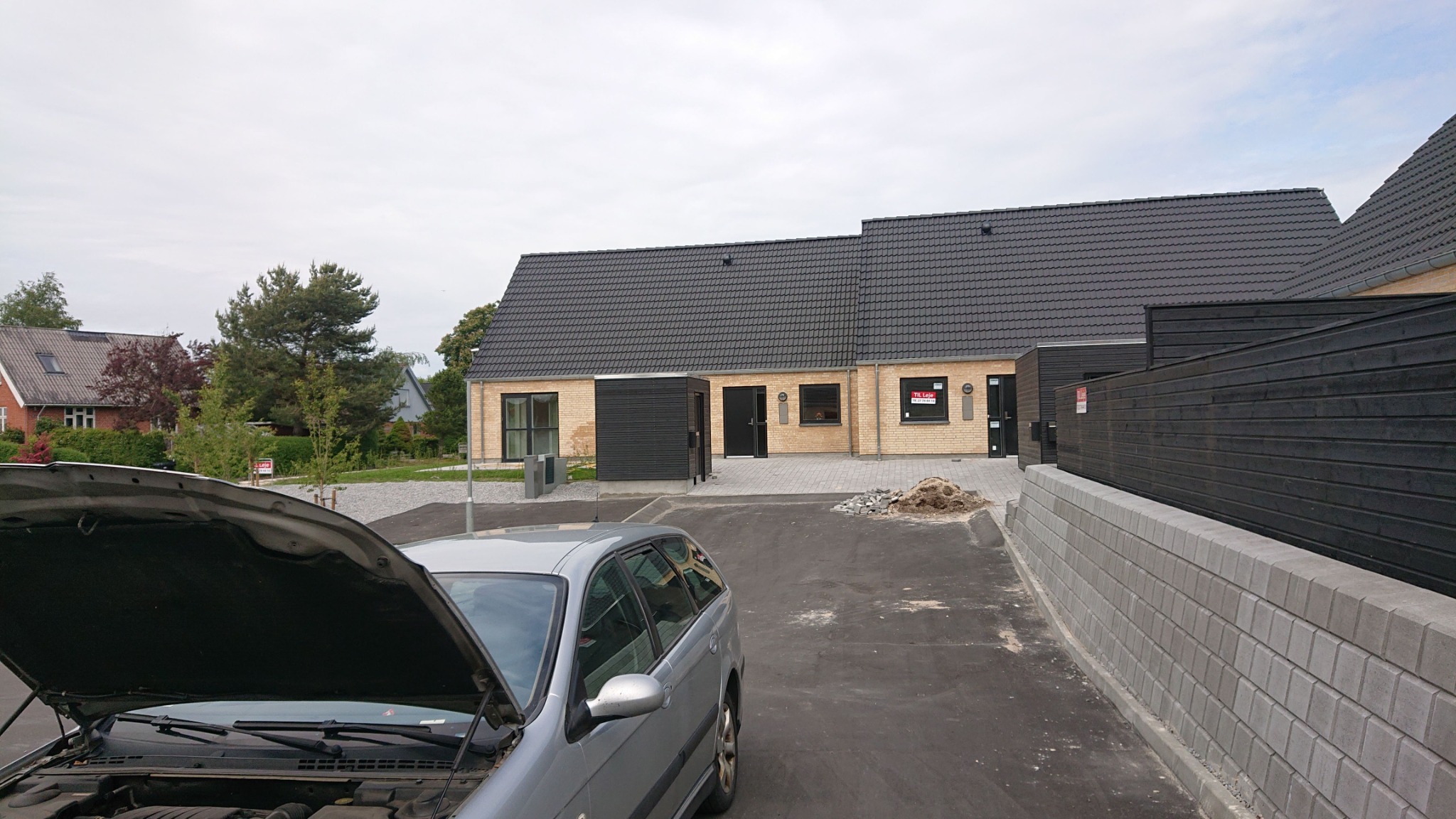 Nowoczesny dom z żółtej cegły z czarnym dachem i elementami wykończenia, widoczny z perspektywy samochodu z otwartą maską na podjeździe.