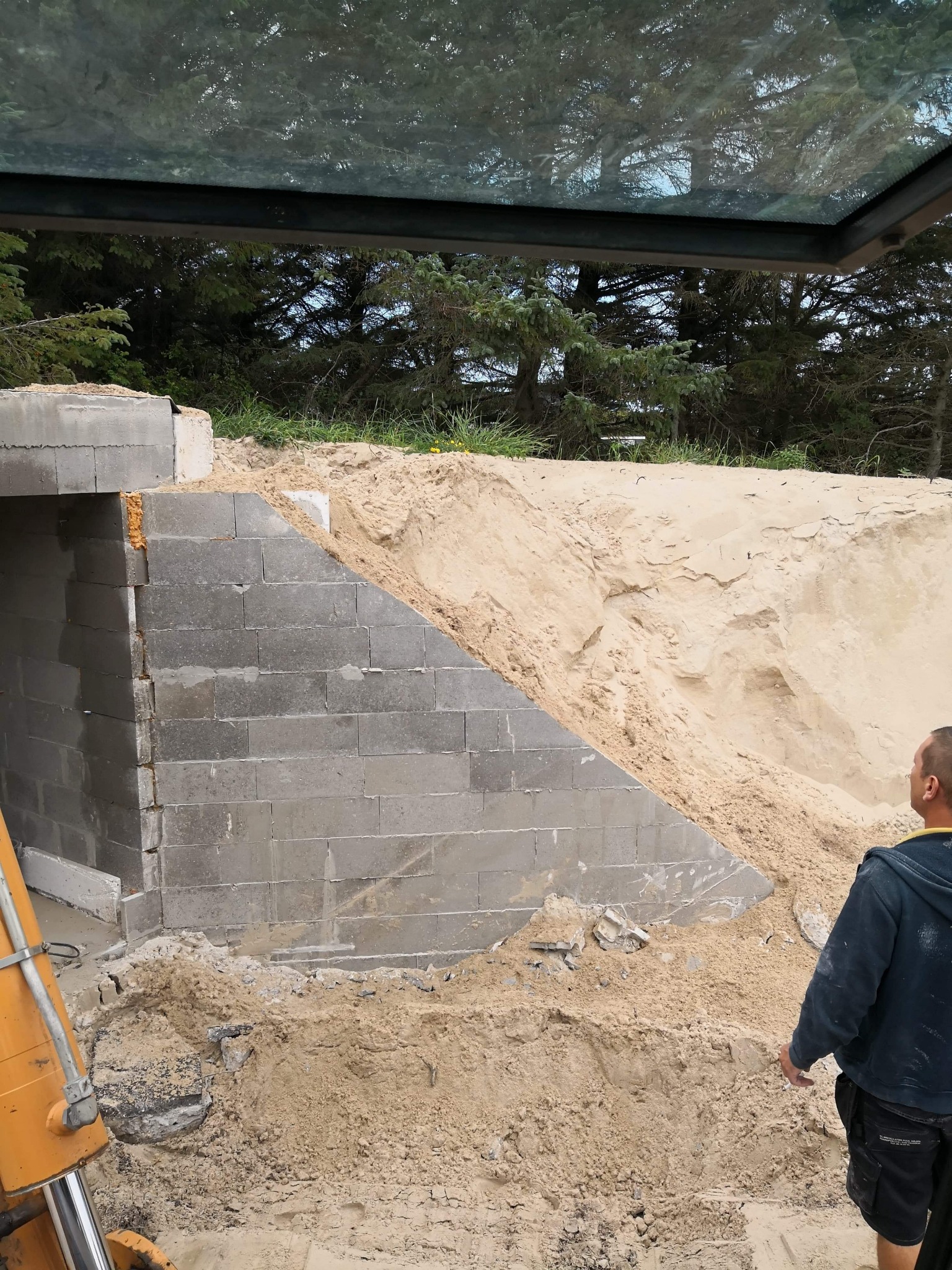 Wznoszenie ściany z szarych bloczków betonowych, częściowo zasypanej piaskiem, widoczne elementy żółtej maszyny budowlanej i fragment sylwetki mężczyzny.
