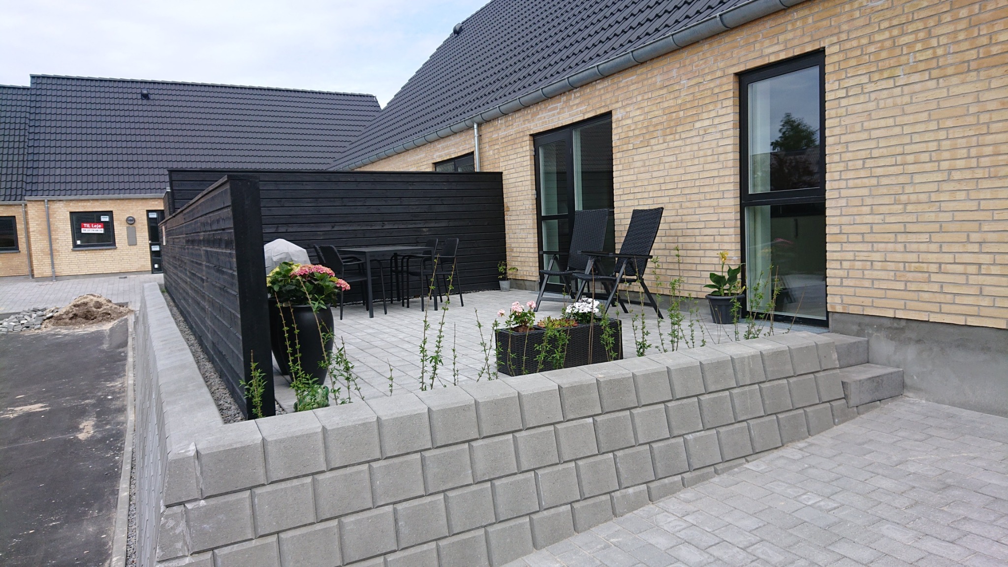 Nowoczesny dom murowany z żółtej cegły z czarnymi ramami okiennymi, tarasem wyłożonym szarą kostką brukową i czarnym drewnianym ogrodzeniem.