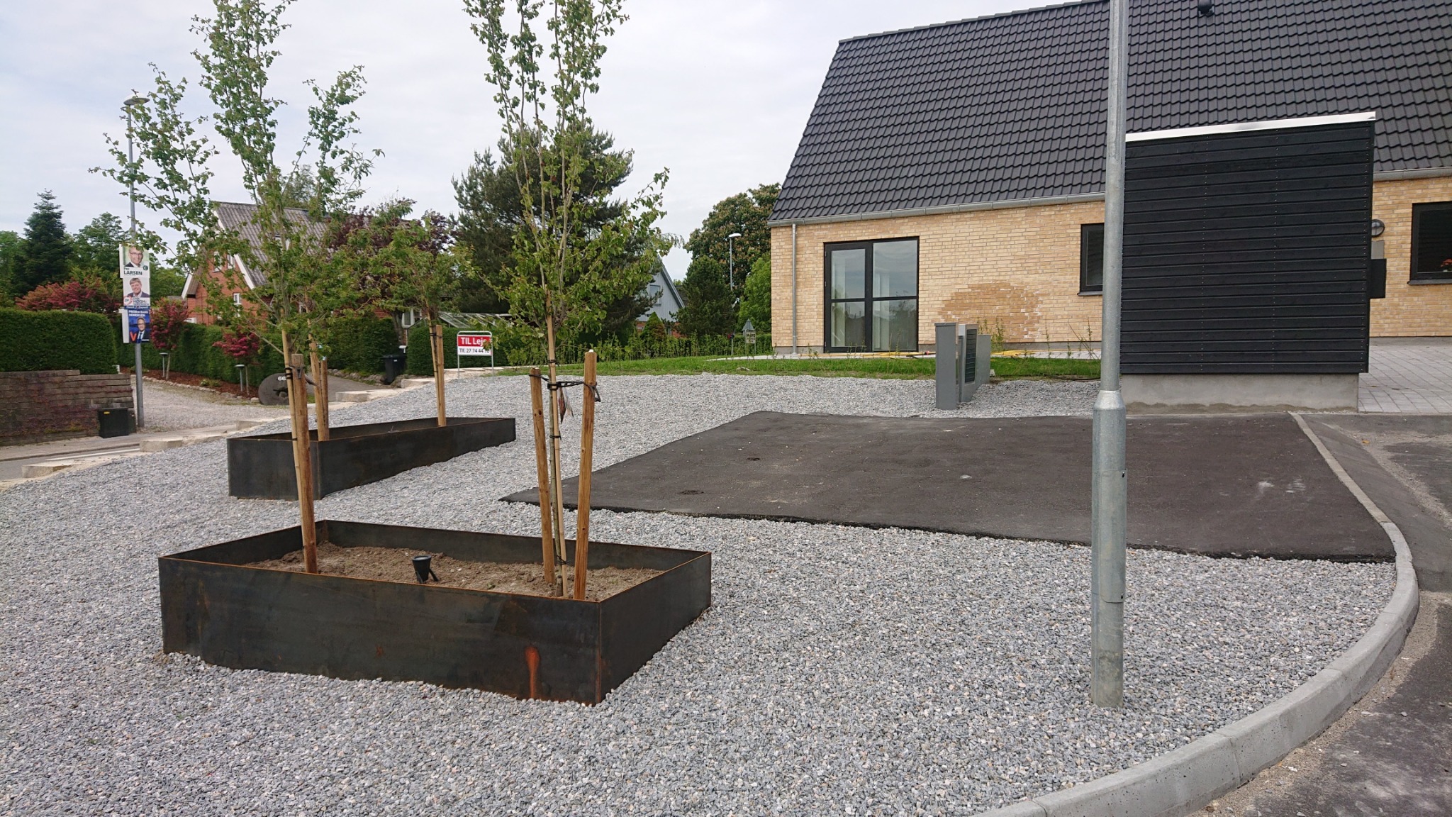 Nowoczesny dom z jasnej cegły z ciemnym dachem i czarną elewacją, otoczony szarym żwirem i metalowymi donicami z młodymi drzewkami.