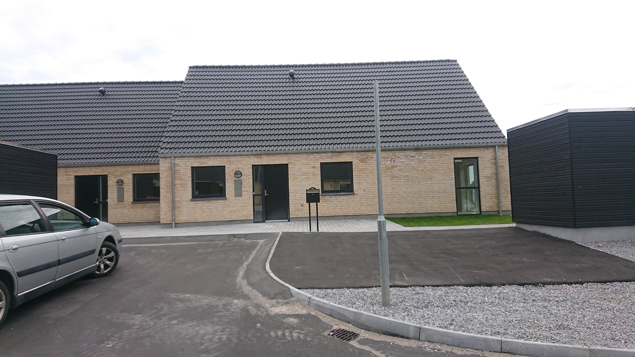 Nowoczesny dom jednorodzinny z jasnej cegły z ciemną dachówką, czarnymi oknami i drzwiami, widok z zewnątrz, szary samochód zaparkowany z boku.