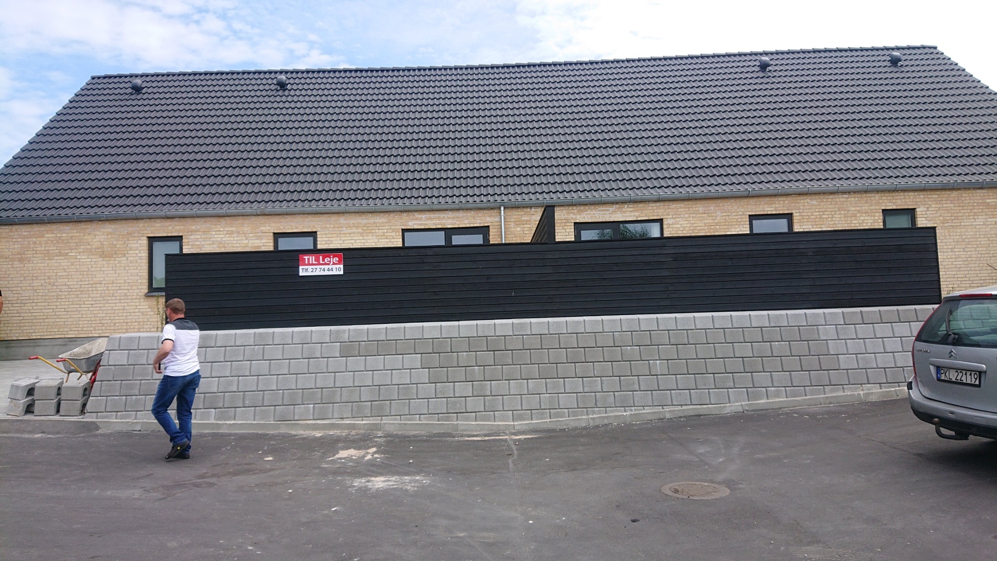 Nowo wybudowany dom z jasnej cegły z ciemną drewnianą ścianą i szarą dachówką, widoczny fragment murku z szarej kostki, w tle błękitne niebo.