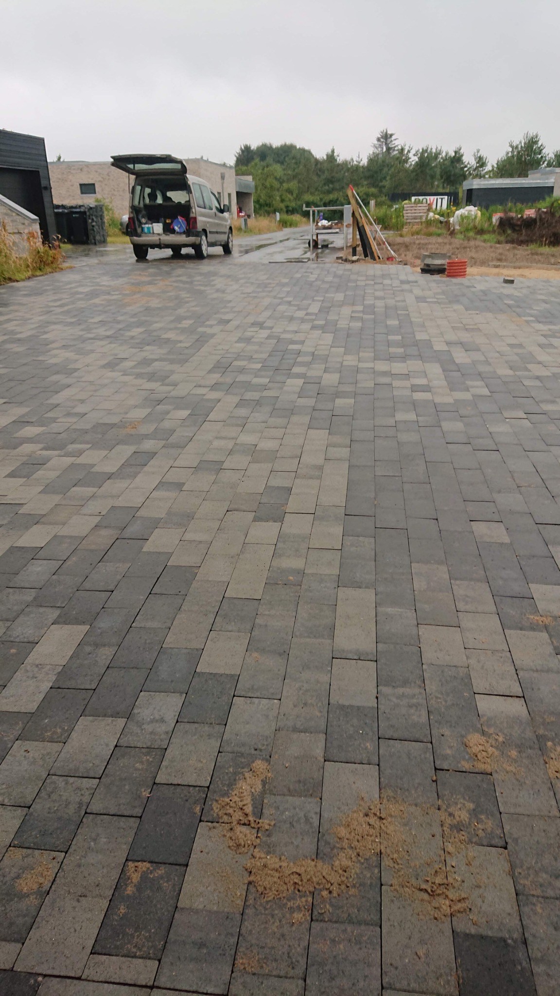 Ułożona kostka brukowa w odcieniach szarości na podjeździe, z widocznym samochodem dostawczym z otwartą klapą bagażnika w tle oraz elementami budowy.