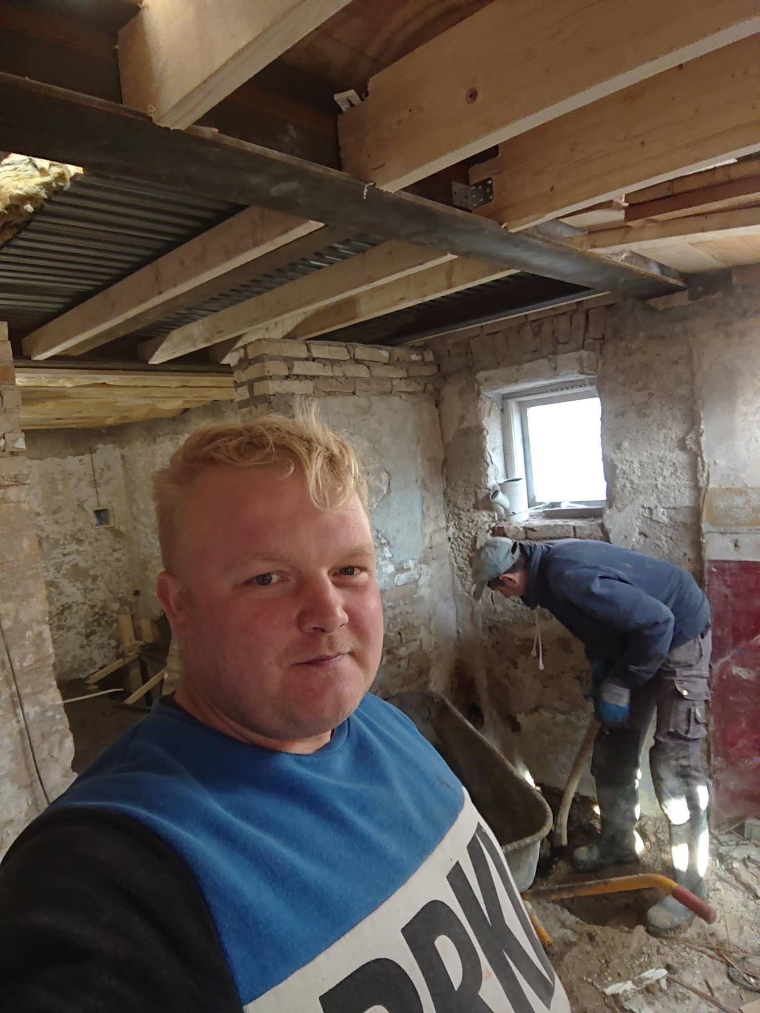 Mężczyzna robiący selfie na tle remontowanego wnętrza z odsłoniętymi cegłami i drewnianą konstrukcją stropu, drugi pracownik w tle zbiera gruz do taczki.