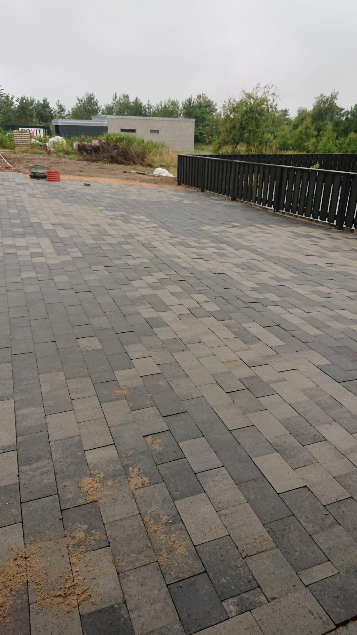 Ułożona kostka brukowa w odcieniach szarości, widoczne ślady piasku, w tle nowoczesny budynek z płaskim dachem i fragment drewnianego ogrodzenia.