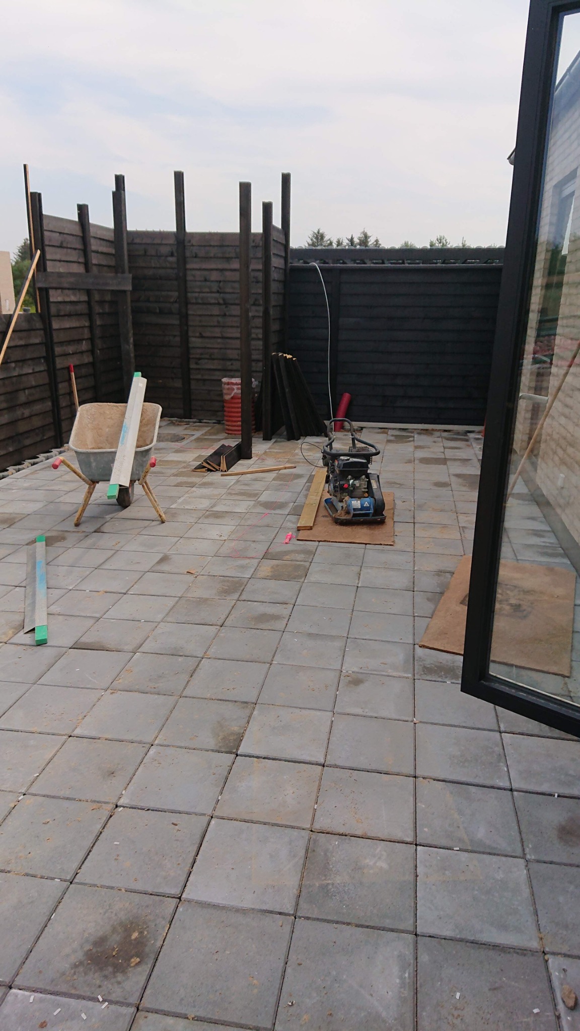 Układanie szarej kostki brukowej na posesji z częściowo ukończonym, ciemnym ogrodzeniem drewnianym; widoczna taczka, poziomica i zagęszczarka w tle.
