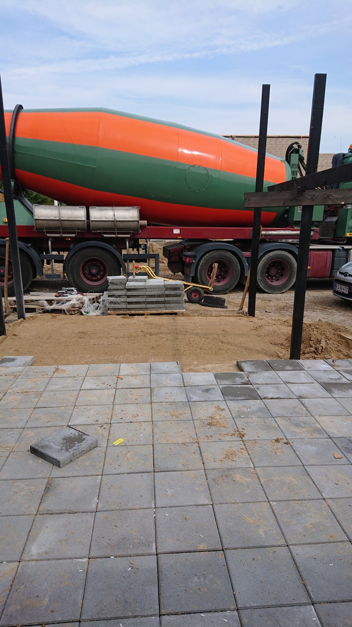 Układanie szarej kostki brukowej na piaskowym podłożu, w tle betoniarka w kolorach pomarańczowo-zielonym oraz sterta kostki brukowej na palecie.