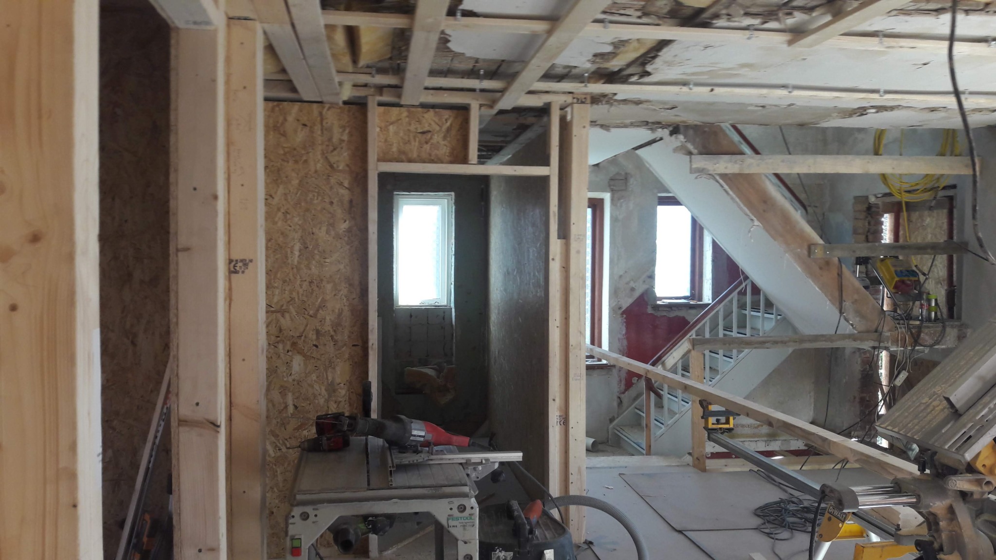 Wnętrze domu w trakcie budowy: widoczne drewniane ramy ścian działowych, fragment schodów, okno, płyty OSB, narzędzia budowlane i resztki starego wykończenia.