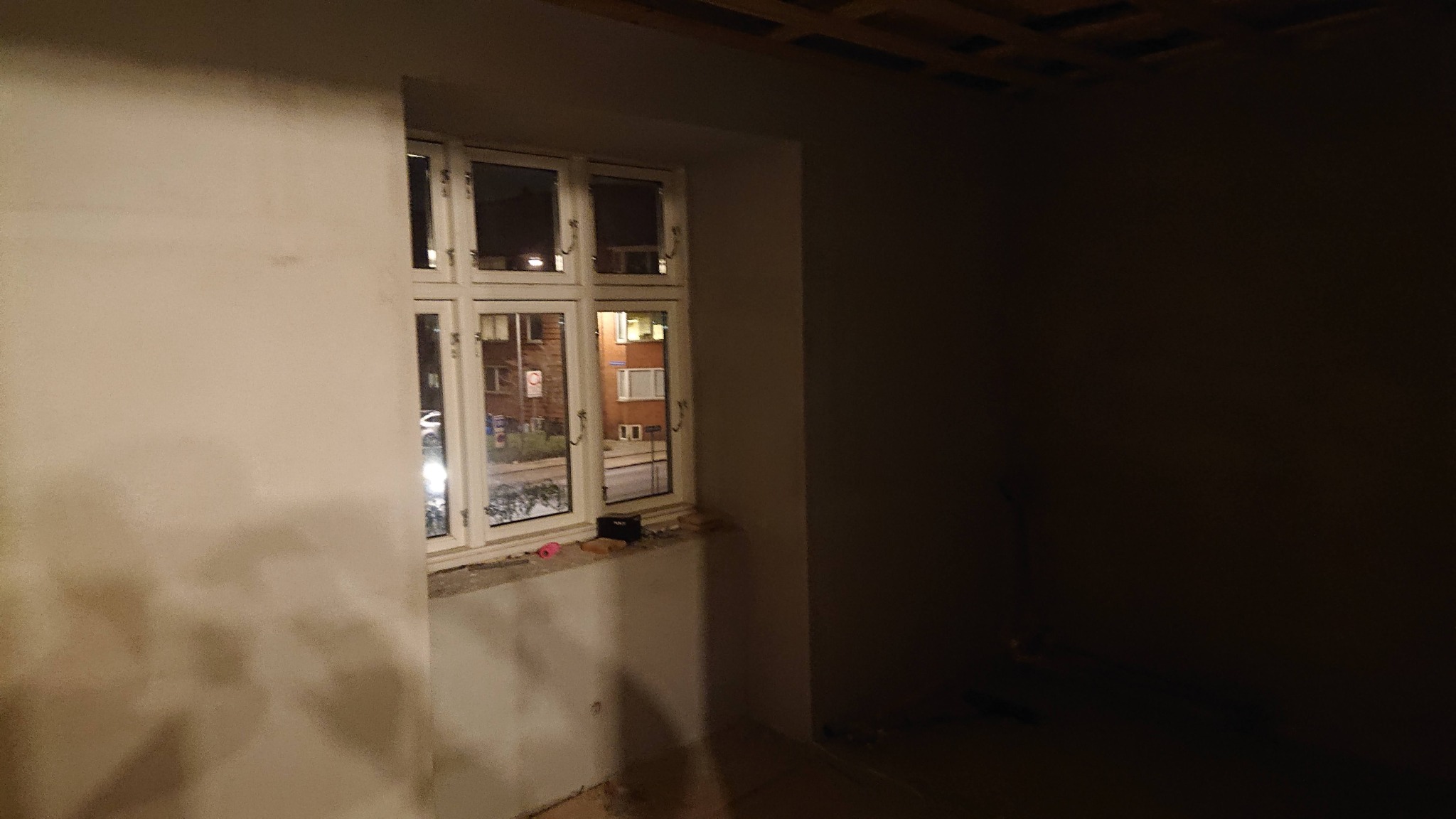 Wnętrze pokoju w trakcie tynkowania ścian, widoczne okno z widokiem na ulicę nocą i surowy sufit.