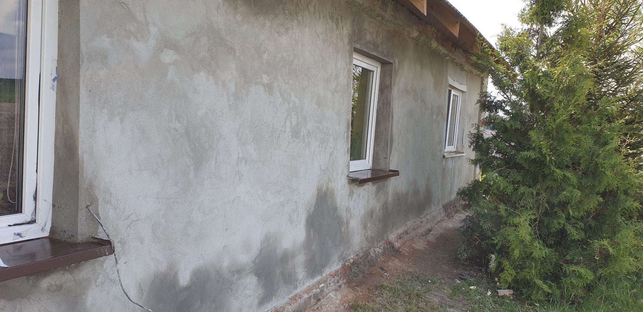 Ściana budynku w trakcie tynkowania maszynowego, widoczne okna z brązowymi parapetami, ślady wilgoci na świeżym tynku i elementy drewniane wystające z dachu.