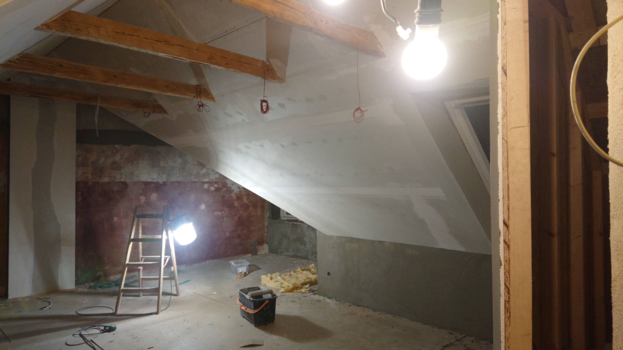 Poddasze w trakcie prac wykończeniowych w Kole: widoczne drewniane belki stropowe, oświetlenie robocze, częściowo położone tynki, drabina i materiały budowlane.