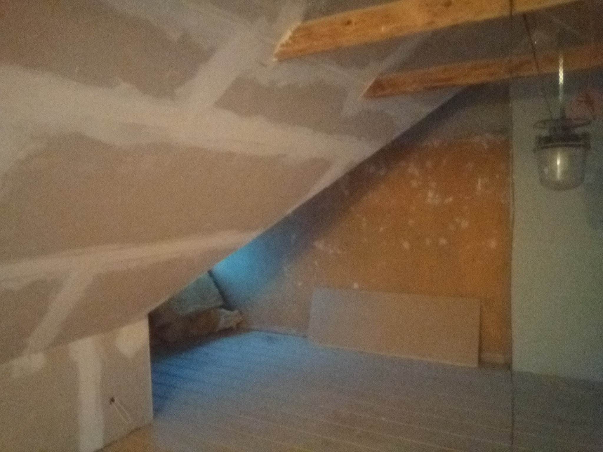 Poddasze w trakcie wykańczania, widoczne płyty gipsowo-kartonowe na skosach sufitu, szpachlowane łączenia, drewniane belki stropowe i surowa podłoga z desek.