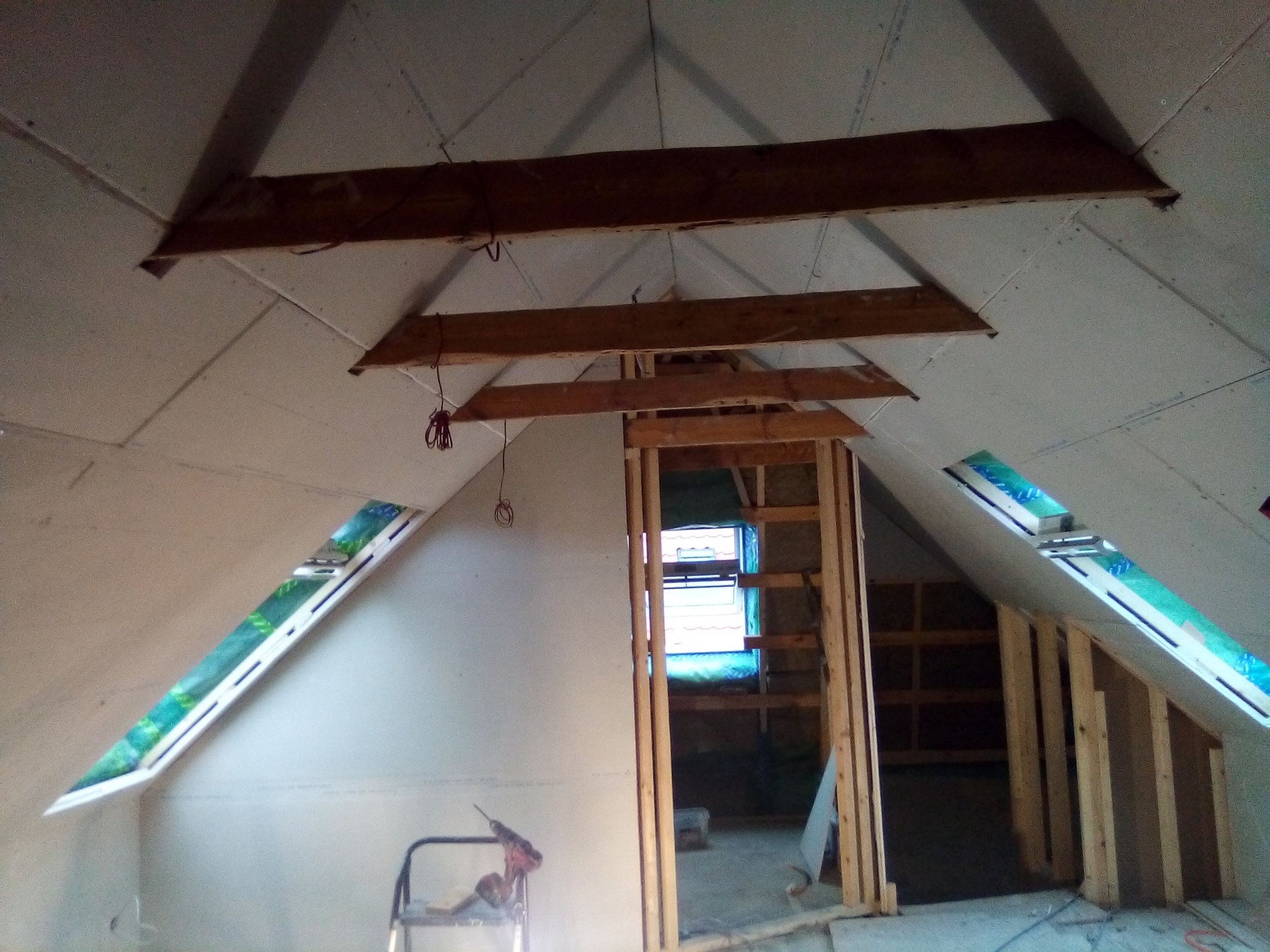 Adaptacja poddasza: widoczne drewniane belki konstrukcyjne, okna dachowe z folią ochronną, ściany pokryte płytami gipsowo-kartonowymi, drabina z wiertarką w tle.