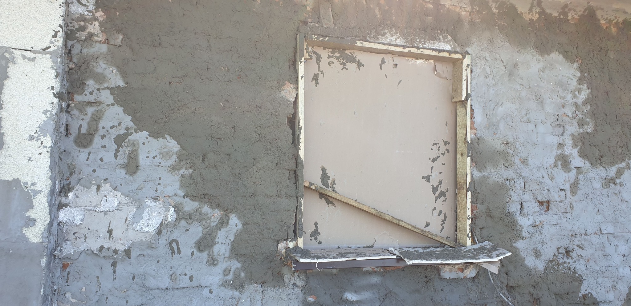 Fragment murowanej ściany w trakcie tynkowania, z widocznym oknem zabezpieczonym płytą i drewnianą listwą, surowy tynk miejscami nie pokrywa cegieł.