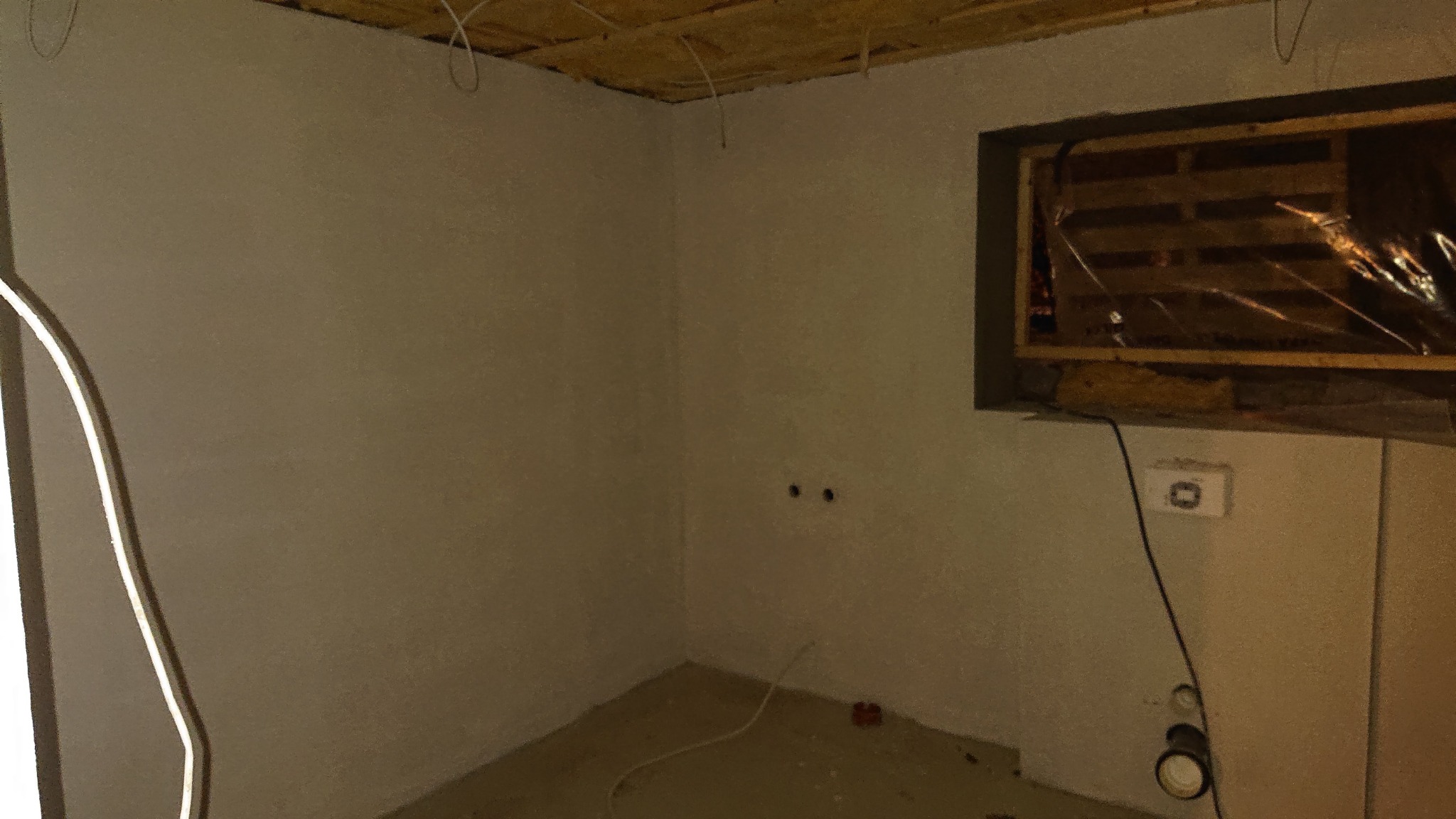 Wnętrze w trakcie prac wykończeniowych: otynkowane ściany, widoczne przewody elektryczne na suficie, zabezpieczone okno folią i drewnianą konstrukcją, puszka elektryczna na ścianie, podłoga...