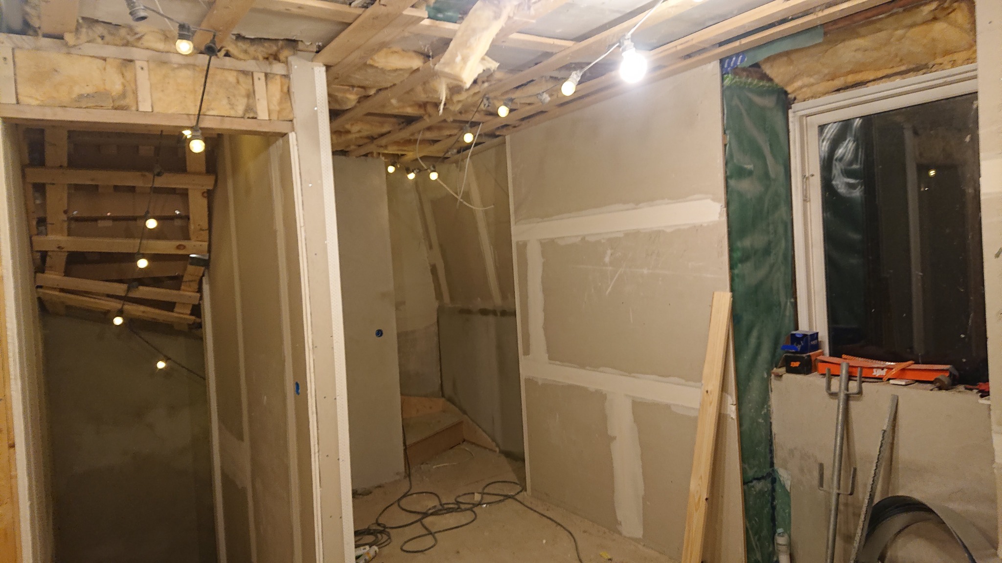 Wnętrze domu w trakcie prac wykończeniowych: widoczne ocieplenie stropu, surowy tynk na ścianach, okno zabezpieczone folią i narzędzia na parapecie, dekoracyjne lampki żarówki.