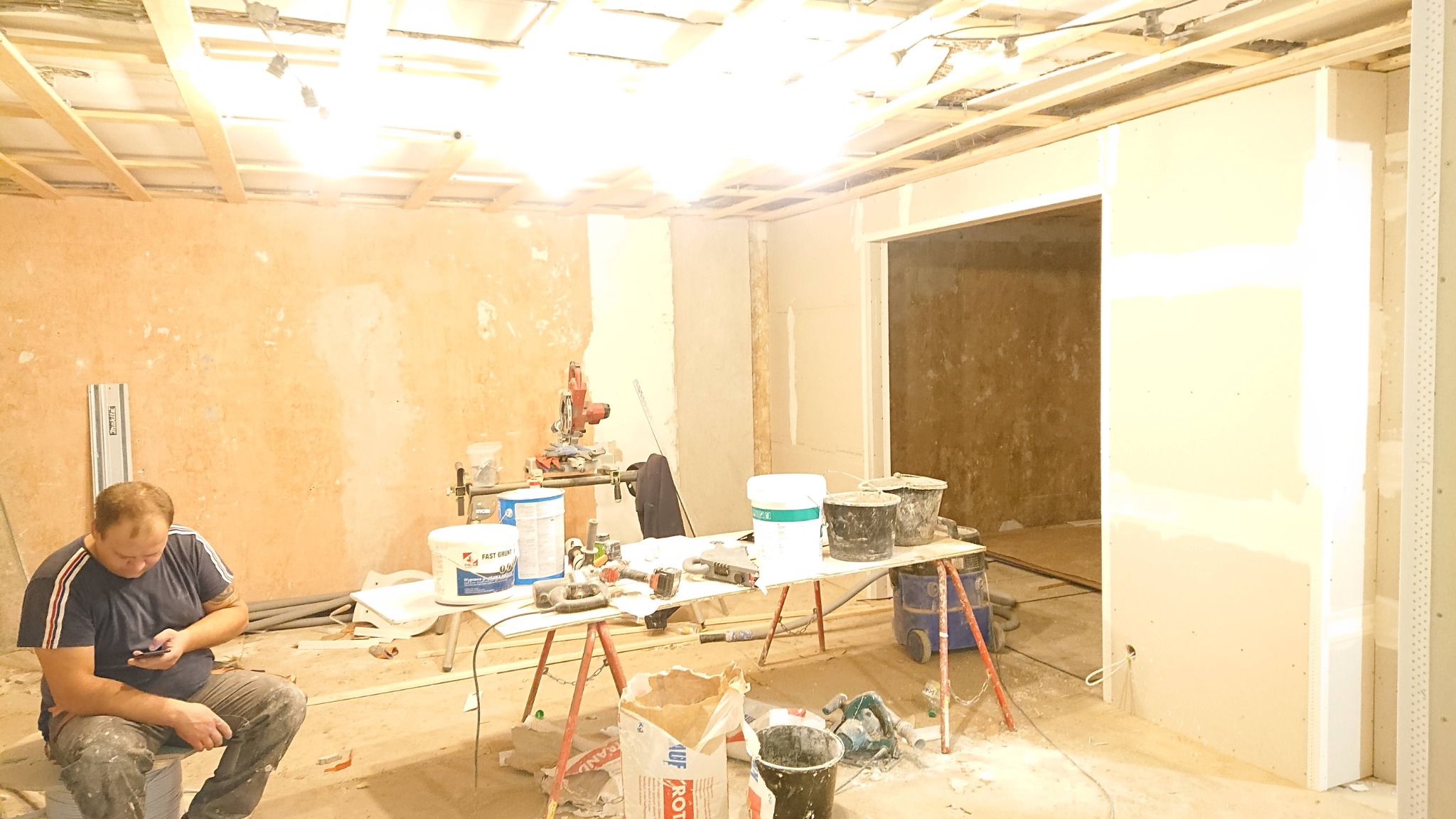 Wnętrze w trakcie remontu: widoczne ściany przygotowane do tynkowania, drewniana konstrukcja sufitu, narzędzia i materiały budowlane rozłożone na stole oraz mężczyzna siedzący na wiadrze...