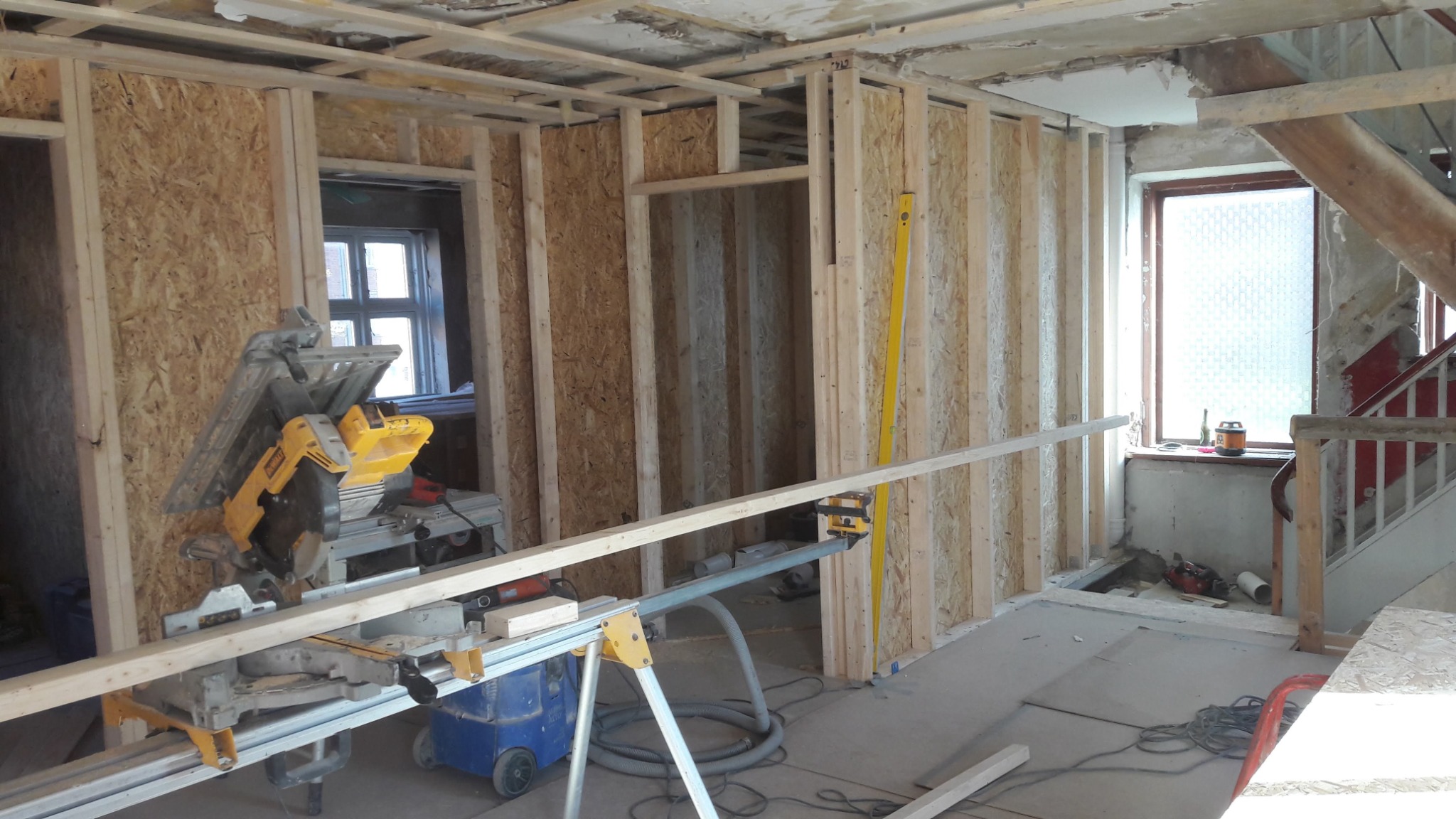 Wnętrze w trakcie budowy: drewniana konstrukcja ścian szkieletowych z płytami OSB, piła stołowa DeWalt na statywie, poziomica, schody w tle.