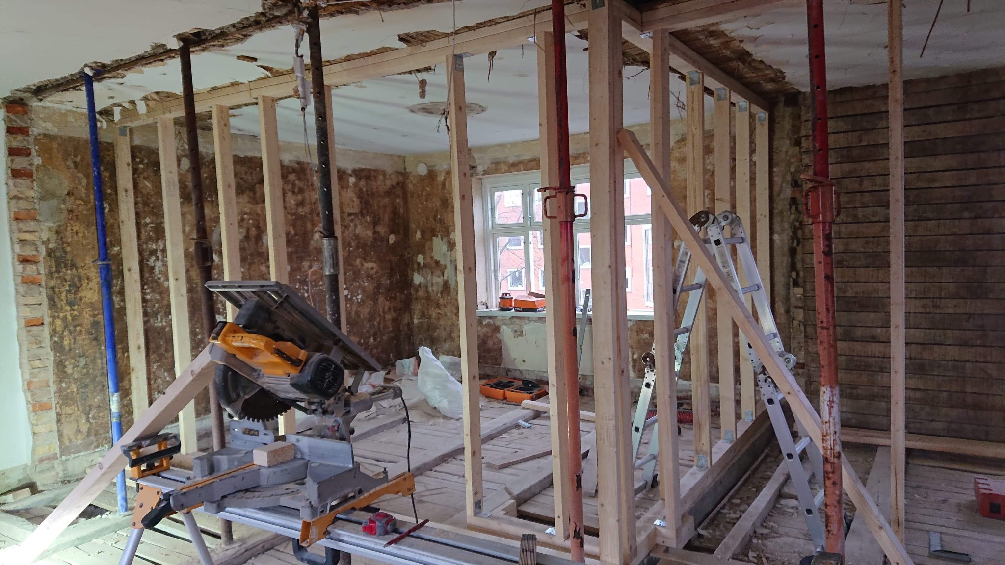 Wnętrze remontowanego domu z odsłoniętymi cegłami, drewnianą konstrukcją ścian działowych i piłą stołową na pierwszym planie, widoczne podpory stropowe i drabina.