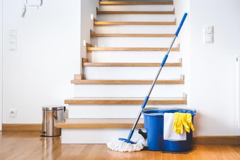 Mop oparty o niebieskie wiadro z rękawicami i ściereczką na tle drewnianych schodów i błyszczącej podłogi.