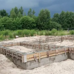 Przygotowany szalunek fundamentów zbrojonych, częściowo zalany betonem, na piaszczystym terenie, z widoczną zielenią w tle i fragmentem pojazdu budowlanego z lewej strony.