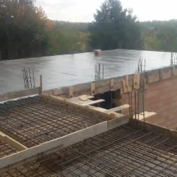 Wylewanie betonu na strop domu z widoczną konstrukcją zbrojeniową i drewnianym szalunkiem, w tle las