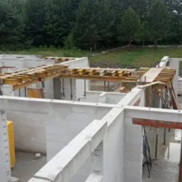 Wznoszenie ścian parteru domu z betonu komórkowego, widoczne żółte opakowania materiałów budowlanych i drewniane szalunki stropu.