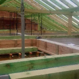 Wnętrze budowy domu w trakcie wznoszenia konstrukcji dachu i szalunk&oacute;w, widoczne drewniane elementy konstrukcyjne impregnowane na zielono, stalowe podpory i częściowo wymurowane ściany z czerwonej...