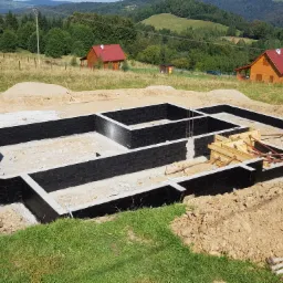 Zalane fundamenty z czarną izolacją przeciwwilgociową, widoczne szalunki i zbrojenia, w tle krajobraz z domami i wzgórzami.