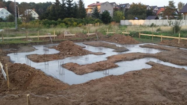 Wylany betonowy fundament z wystającymi prętami zbrojeniowymi pod budowę domu jednorodzinnego, widoczne drewniane szalunki i pryzmy ziemi.