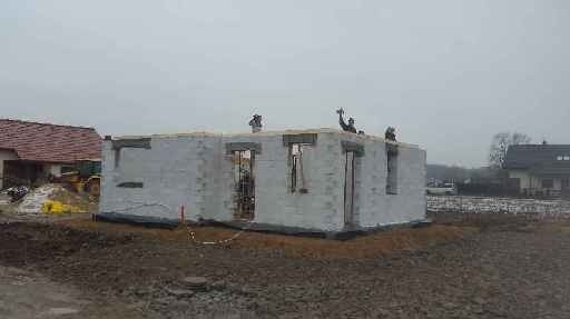 Surowy stan budynku z białych bloczków z widocznymi otworami okiennymi i drzwiowymi, ekipa budowlana na dachu, w tle krajobraz z innymi budynkami i sprzętem budowlanym.