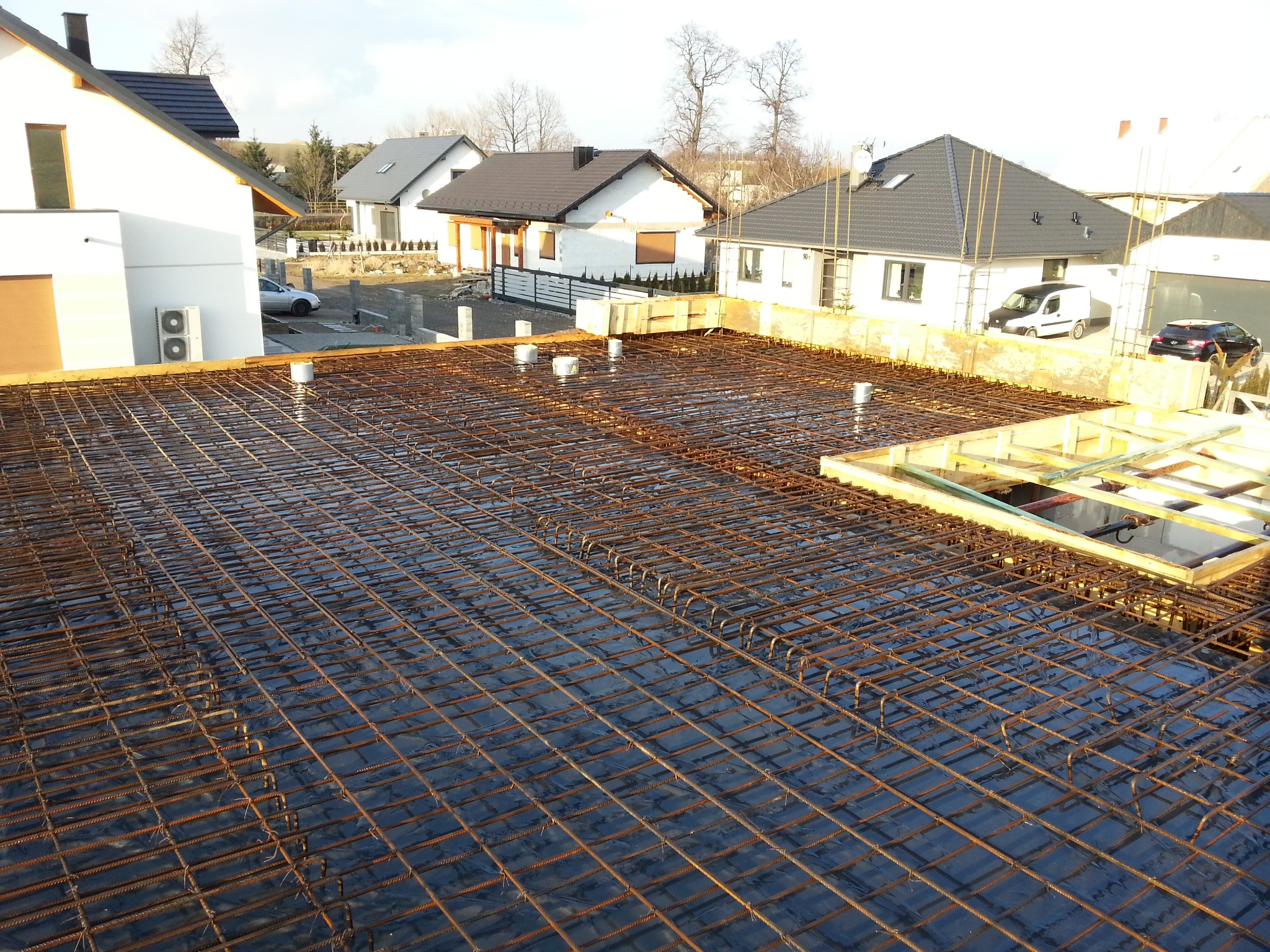 Zbrojenie stropu na folii hydroizolacyjnej, widoczne elementy szalunku i zabudowa osiedla domów jednorodzinnych w tle.