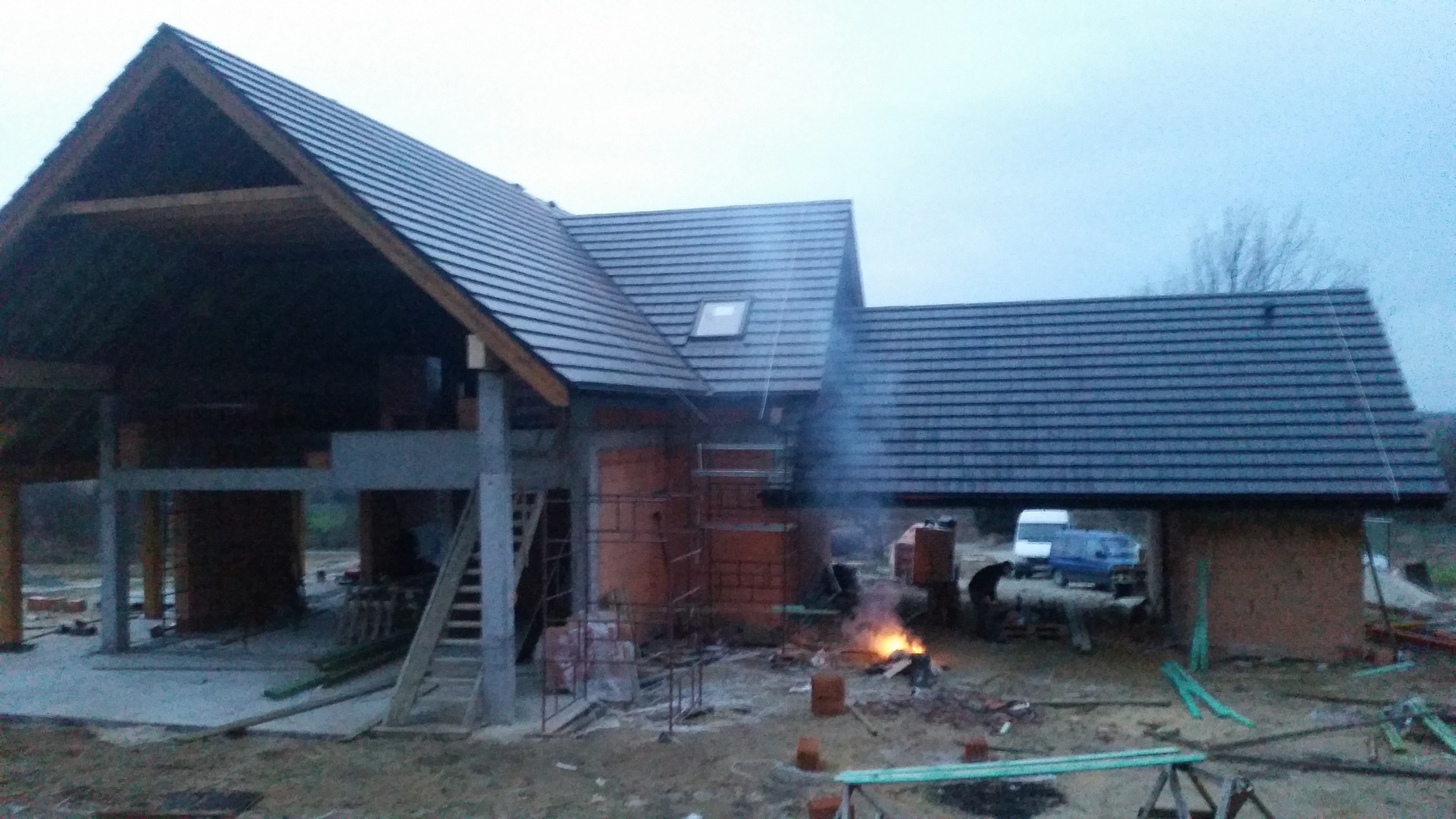 Surowy stan domu murowanego z widoczną konstrukcją dachu, ceglanymi ścianami, rusztowaniami i paleniskiem na zewnątrz.