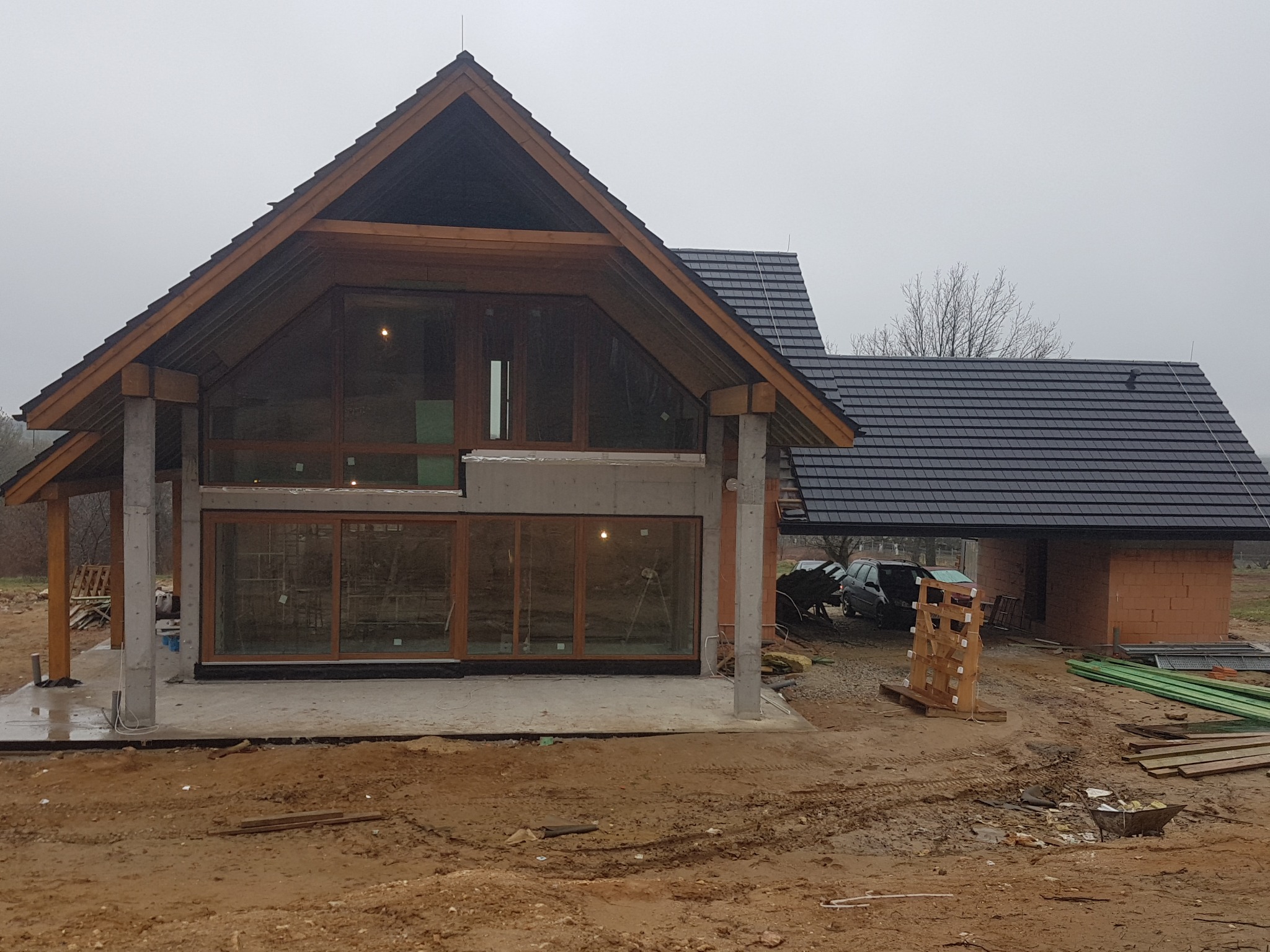 Surowy stan domu z dużymi oknami i drewnianą konstrukcją dachu, widoczny plac budowy z materiałami i błotem.
