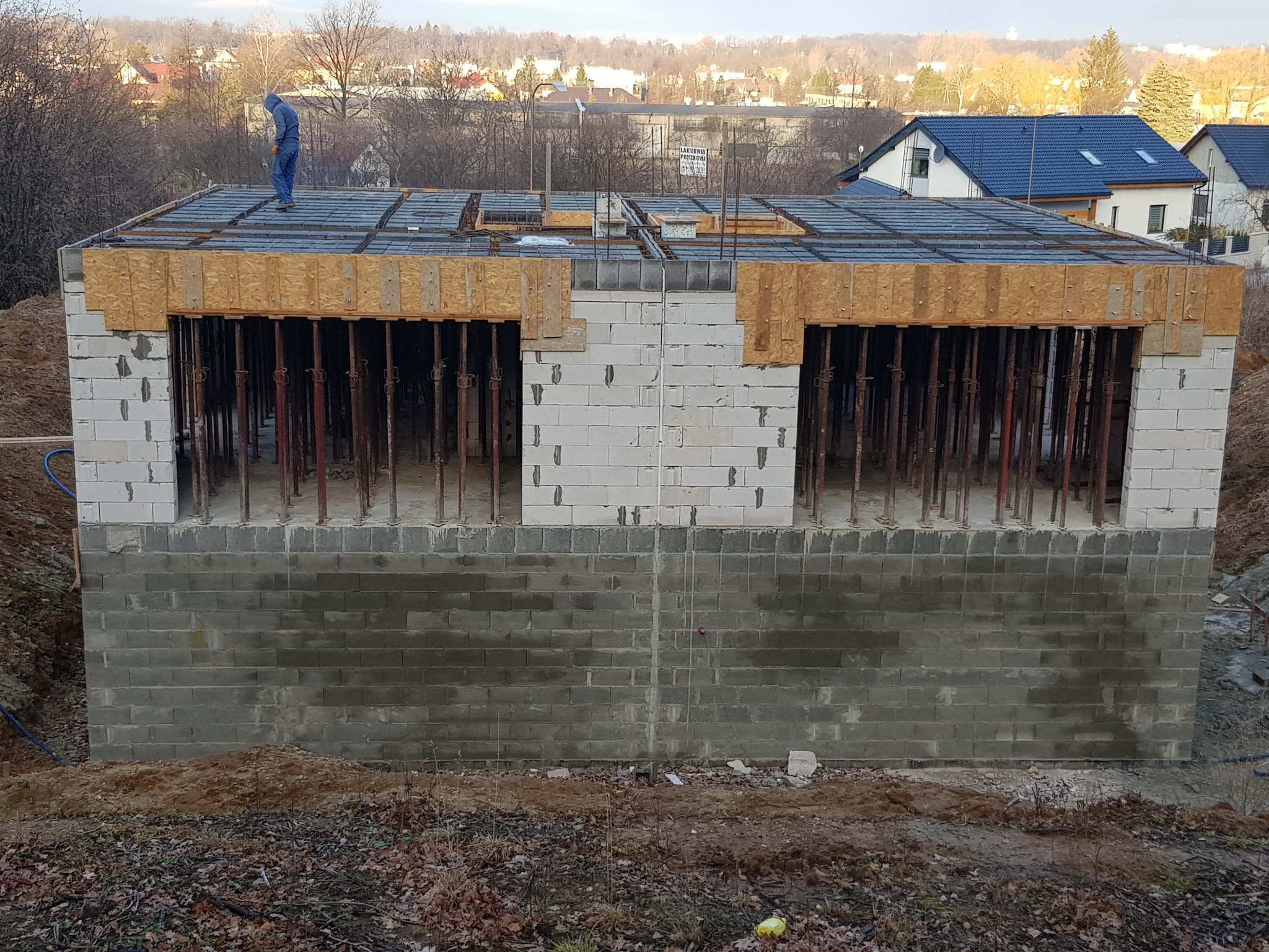 Budowa domu w stanie surowym otwartym: wzniesione ściany fundamentowe z bloczków betonowych, ściany parteru z białych bloczków, drewniane szalunki na stropie, robotnik w niebieskim kombinezonie...