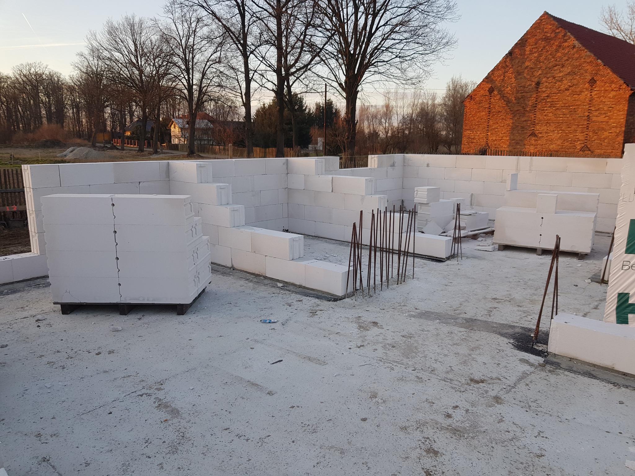 Budowa domu z białych bloczków w Świdnicy, widoczne zbrojenia i paleta z materiałem budowlanym, w tle budynek gospodarczy z czerwonej cegły.