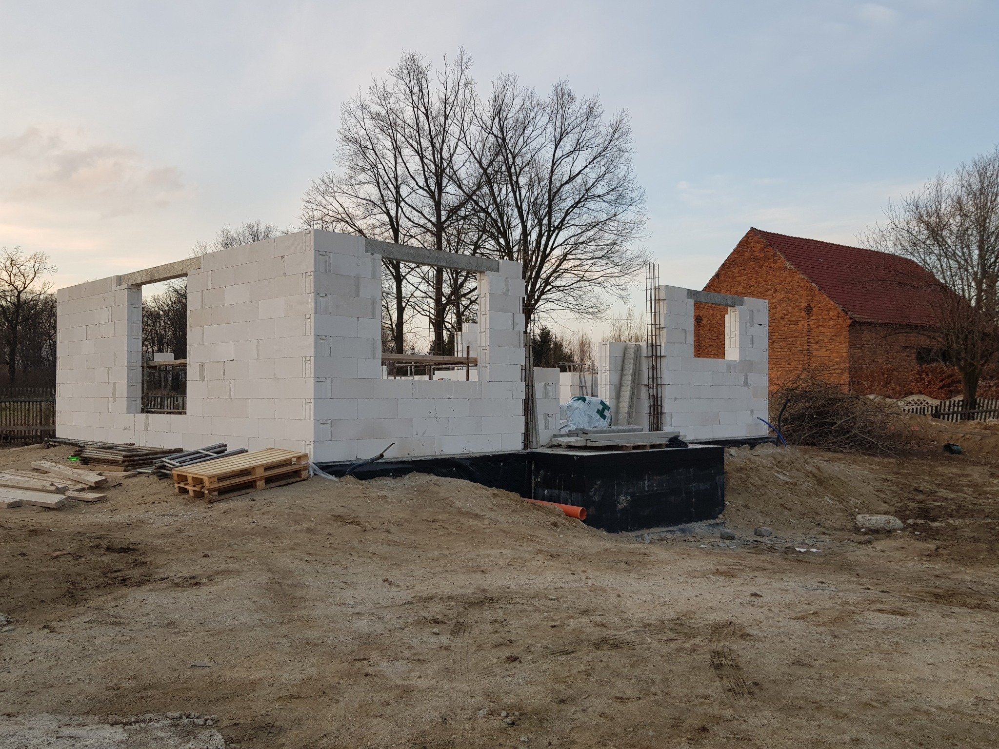 Wznoszone ściany parteru domu z betonu komórkowego, widoczne zbrojenia pionowe, paleta z materiałami budowlanymi na placu budowy, w tle budynek gospodarczy z czerwonej cegły i drzewa bez liści.