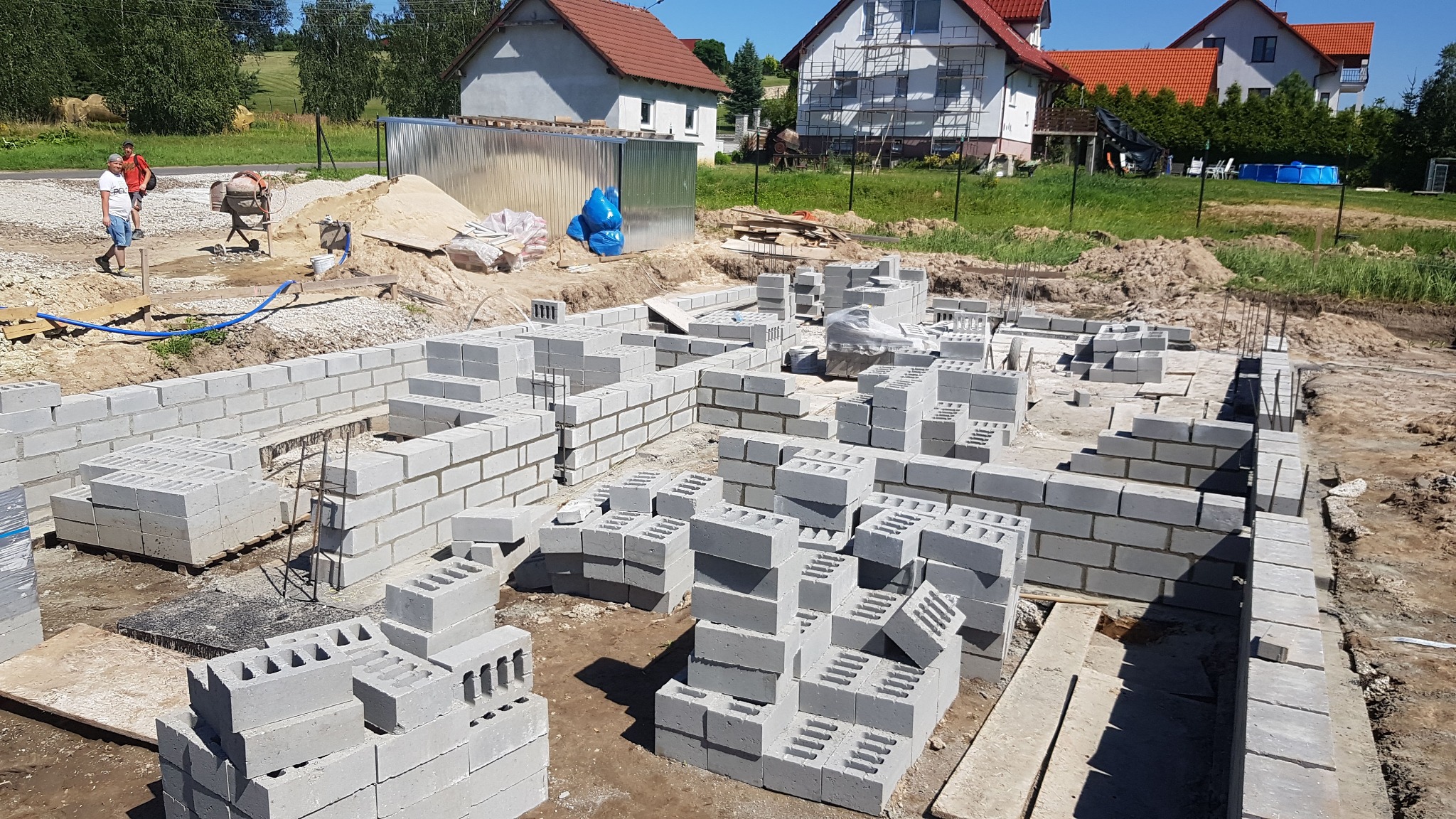 Wznoszenie fundamentów z bloczków betonowych na placu budowy, widoczna betoniarka i dwóch pracowników w tle, domy w zabudowie jednorodzinnej w tle.