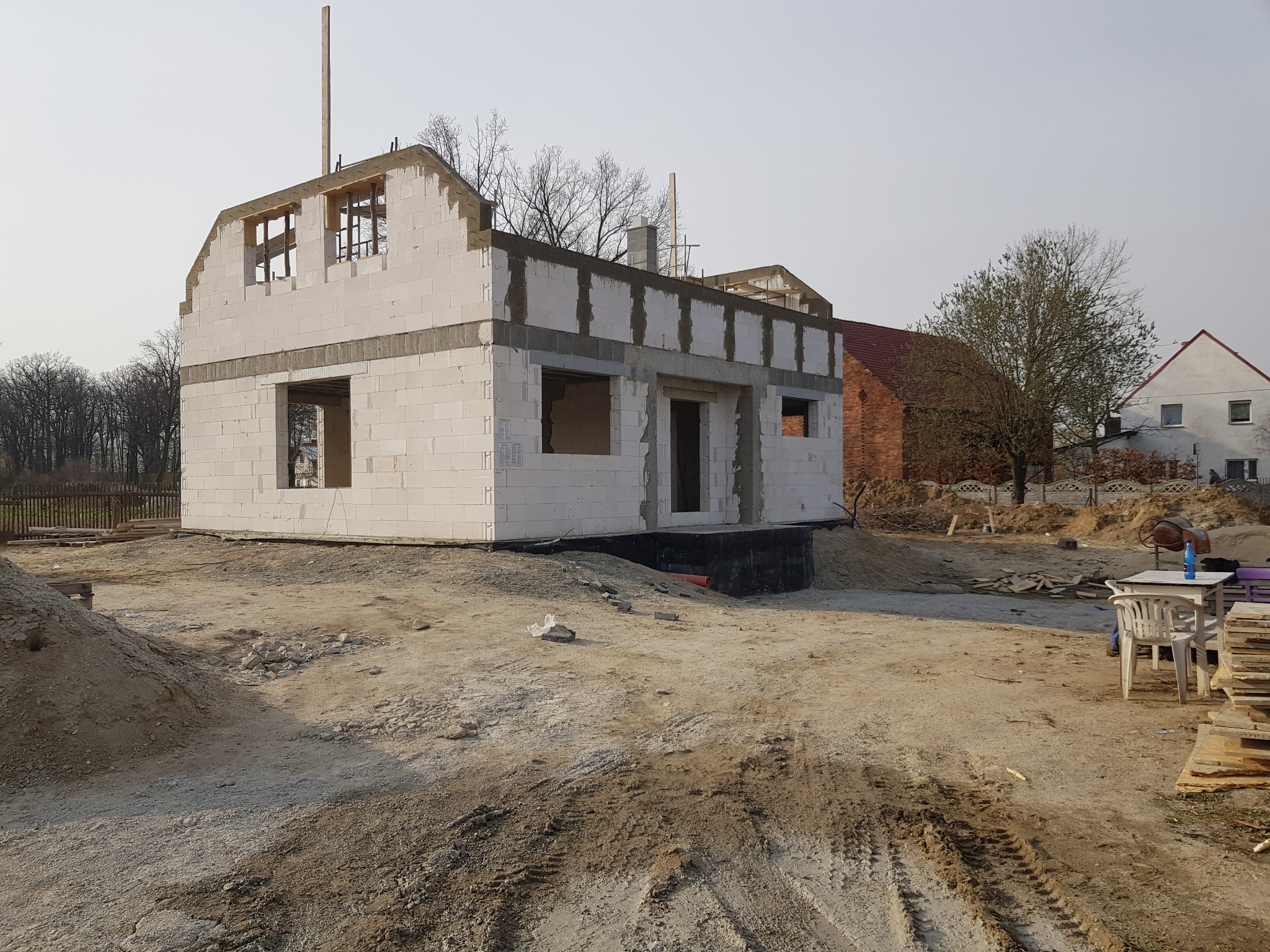 Surowy stan domu murowanego z betonu komórkowego, widoczne ściany parteru i poddasza, zbrojenie wieńca, teren budowy z hałdą piasku i zapleczem socjalnym.