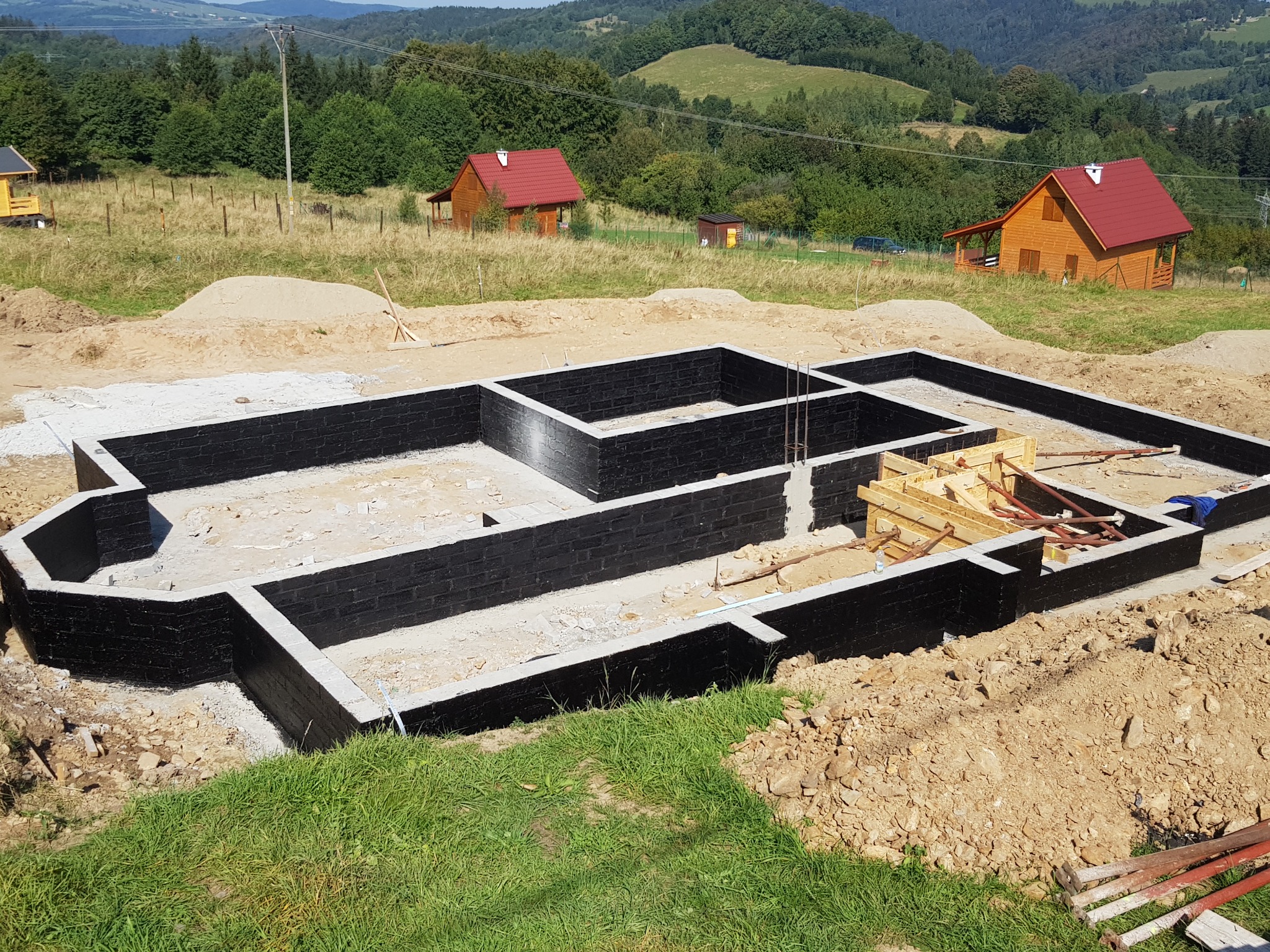 Zalane fundamenty z czarną izolacją przeciwwilgociową, widoczne szalunki i zbrojenia, w tle krajobraz z domami i wzgórzami.