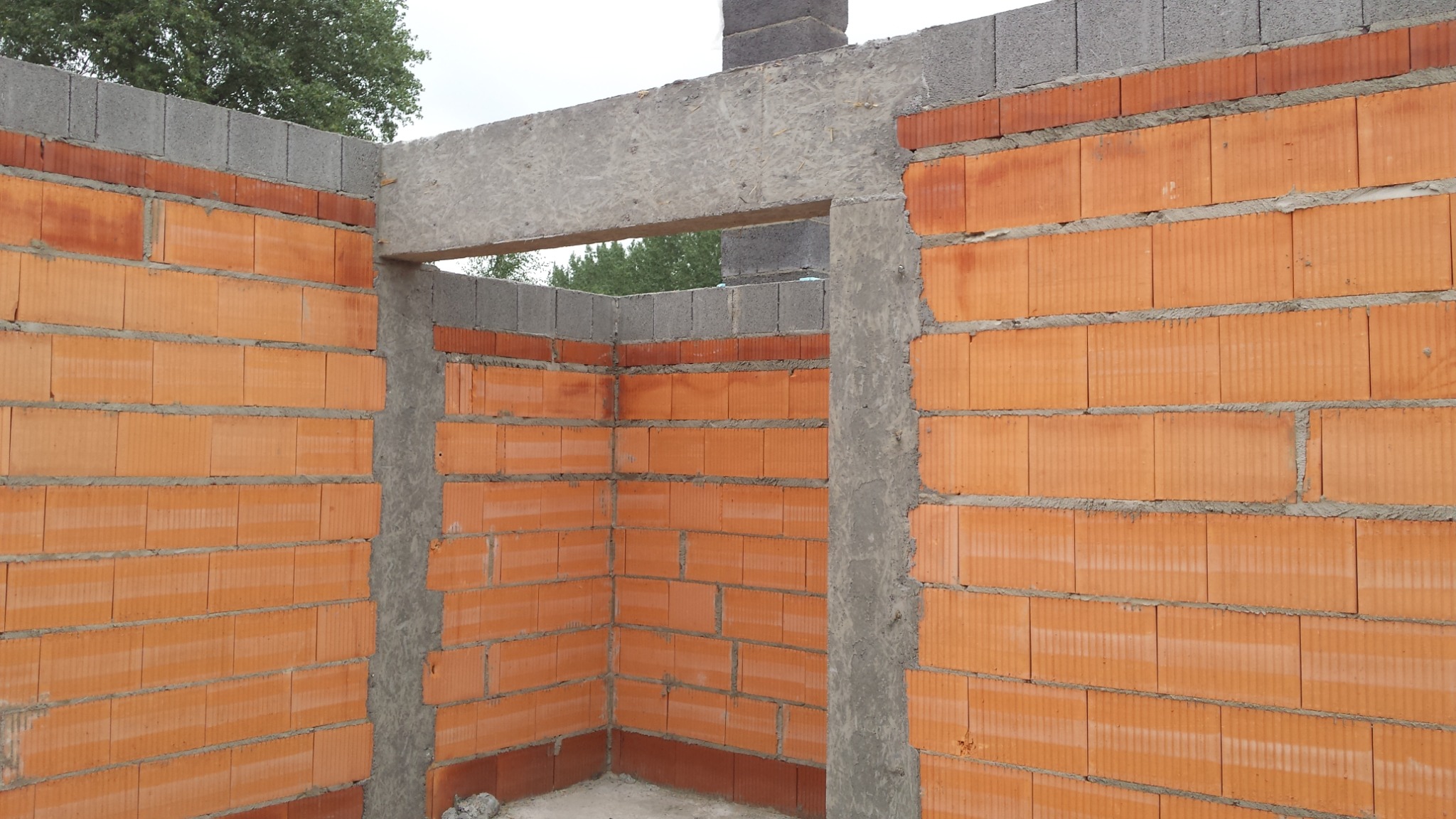 Wewnętrzny róg budynku w trakcie wznoszenia murów z czerwonej cegły kratówki, widoczne betonowe nadproże i szare bloczki na szczycie ścian.