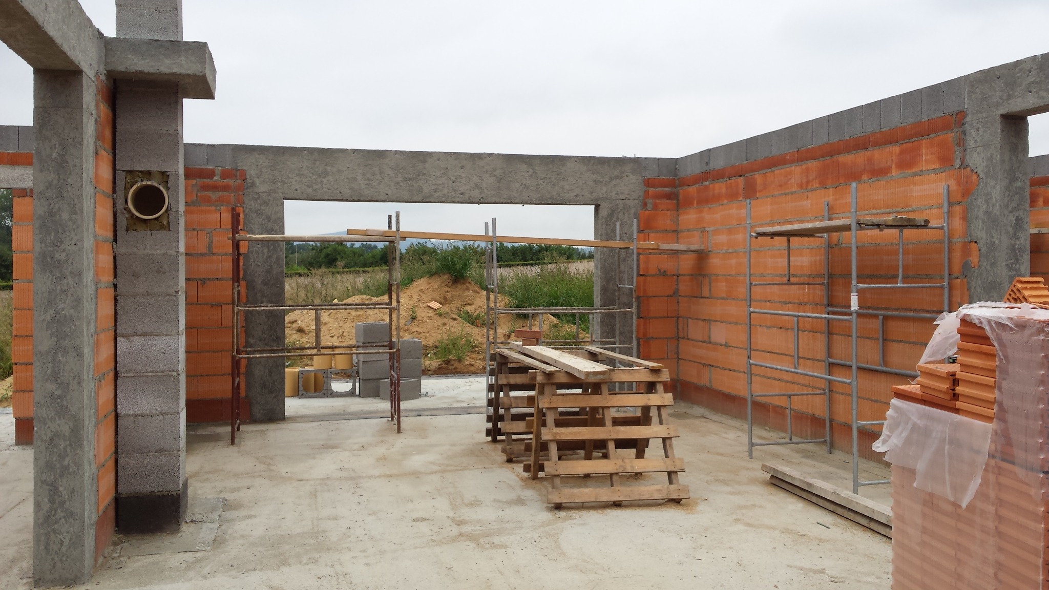 Wnętrze surowego budynku w trakcie wznoszenia murów z czerwonej cegły, widoczne szare betonowe słupy, otwór okienny z widokiem na krajobraz, metalowe rusztowania i palety z cegłami.