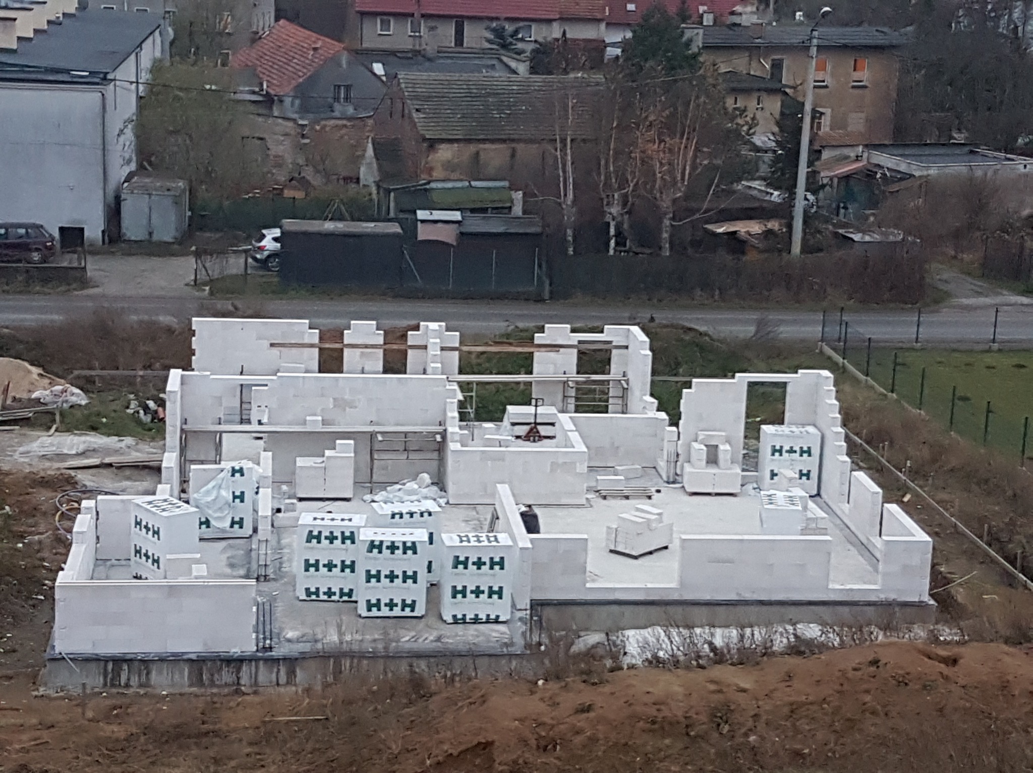 Wznoszenie ścian parteru domu z bloczków betonu komórkowego H+H, widok z góry na plac budowy i okoliczne zabudowania w tle.