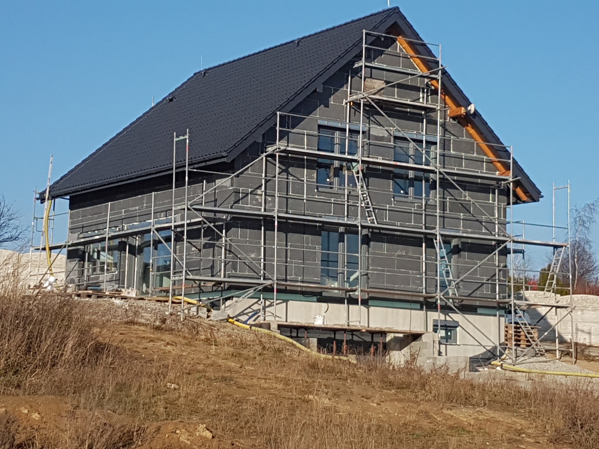 Dom w trakcie budowy, ocieplony styropianem, z czarnym dachem, otoczony rusztowaniem na tle błękitnego nieba.