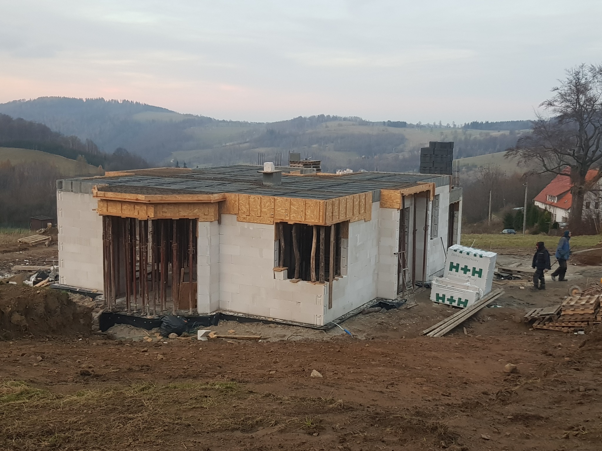 Surowy stan domu z betonu komórkowego w trakcie budowy, widoczne pustaki H+H, drewniane szalunki okienne, krajobraz pagórkowaty w tle.